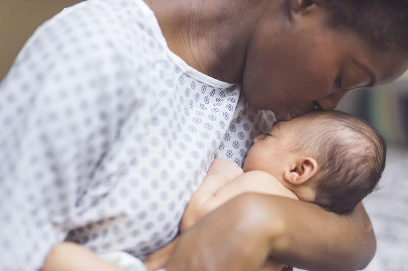 A photo of a woman wearing a hospital gown, holding her newborn baby and kissing their head, which has a flat spot. 