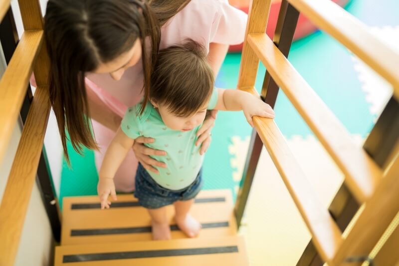 A photo of a woman wearing pink scrubs assisting a child walking up wooden steps. 